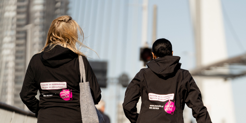 Twee productieleiders van Mooi Echt! lopen over de Erasmusbrug tijdens een fotoshoot in Rotterdam.